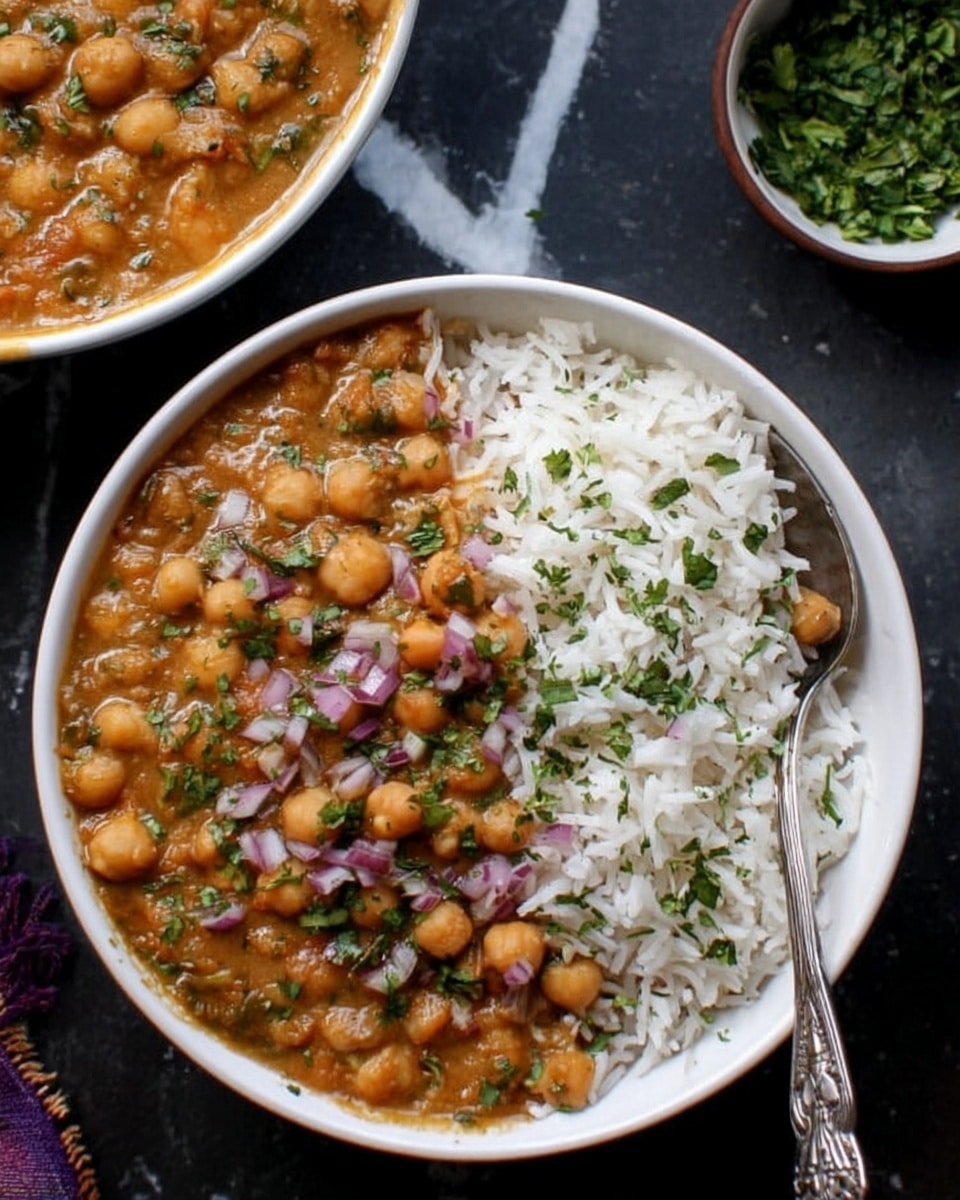 A white bowl filled with two layers: on the left side, fluffy white rice sprinkled with green chopped herbs, and on the right side, a thick brown chickpea curry with a creamy texture, dotted with chickpeas and tiny pieces of red onion and herbs on top. A spoon is placed inside the bowl on the right side. The bowl sits on a dark surface with a white marbled texture, with part of another bowl containing the same chickpea curry visible at the top left corner, and a small bowl with green herbs at the top right. Photo taken with an iphone --ar 4:5 --v 7