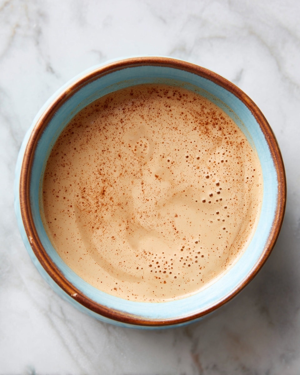 A round bowl with a light blue inside and brown rim is filled almost to the top with a smooth, creamy sauce that is a light beige color with small, evenly spread specks of darker brown. The sauce has a slightly glossy surface and a few tiny bubbles scattered across it. The bowl sits on a white marbled surface, creating a clean and simple background. photo taken with an iphone --ar 4:5 --v 7