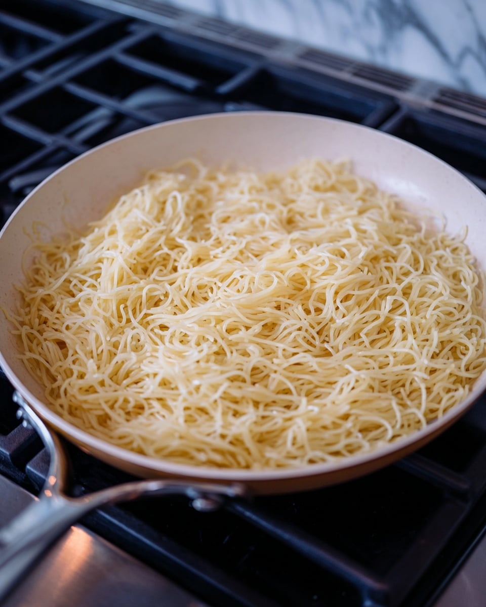 A single layer of thin, pale yellow noodles fills a white pan placed on a dark stove, the noodles appearing soft and tangled with a slightly glossy texture. The pan is round and shallow, with a metal handle extending out on the left side. The background shows part of the stove and a blurry bit of countertop with a white marbled texture. Photo taken with an iphone --ar 4:5 --v 7