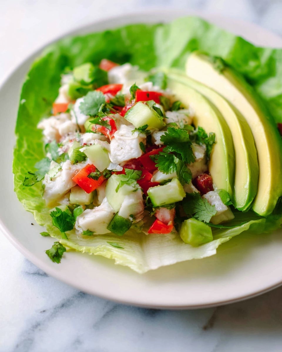 A white plate with a single large green lettuce leaf as the base holds a colorful mixture of small white chunks of cooked fish, bright red diced tomatoes, light green cucumber pieces, and fresh green cilantro leaves scattered evenly across the plate. On top of these ingredients, there are two slices of yellow-green avocado placed on the right side. The plate sits on a white marbled surface, and the overall look is fresh and vibrant. photo taken with an iphone --ar 4:5 --v 7