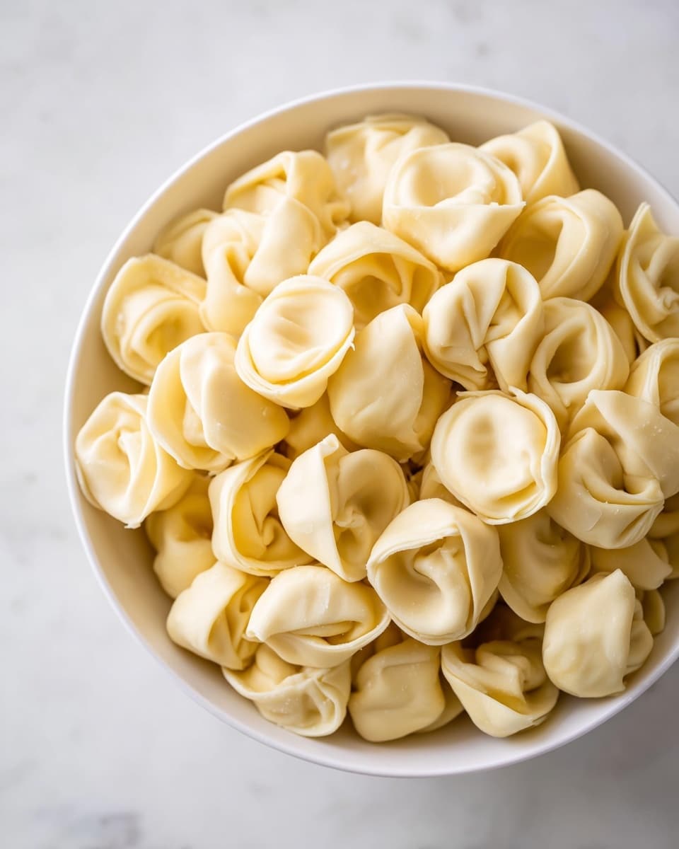 A white bowl filled with many small tortellini pasta pieces stacked closely together, each tortellini showing a smooth pale yellow dough with small folds and curls giving a rounded, stuffed look. The background and surface have a white marbled texture, creating a clean and simple setting for the pasta. photo taken with an iphone --ar 4:5 --v 7