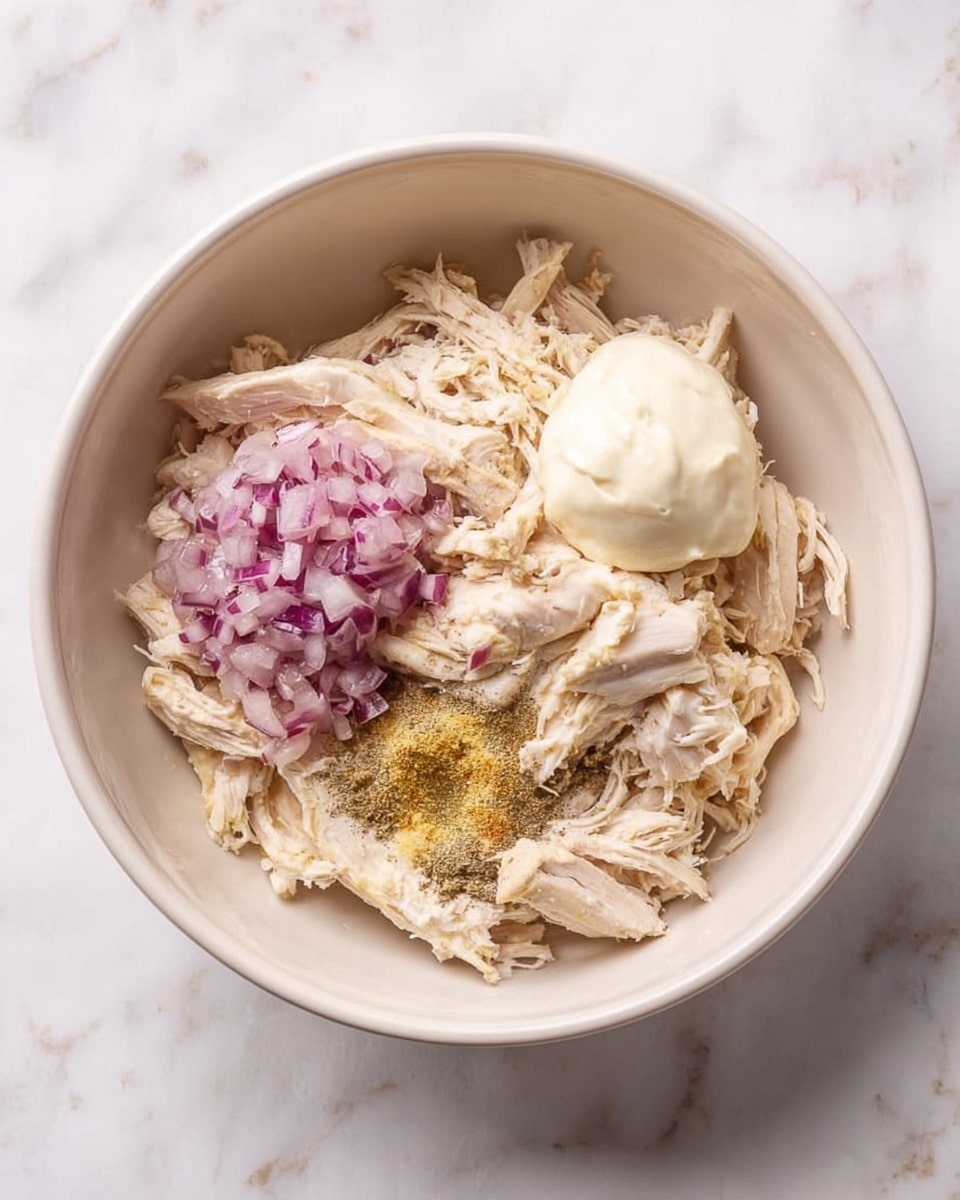 Inside a large white bowl, shredded cooked chicken forms the base layer, light beige in color with a fibrous texture. On top, a small mound of finely chopped red onions adds a purple hue and rough texture. On one side, two dollops of thick creamy substances, one smooth off-white and the other a slightly lighter cream, rest on the chicken. Scattered on the chicken near the onions are seasoning powders in light yellow and dark brown shades. All of this is set against a white marbled surface. photo taken with an iphone --ar 4:5 --v 7