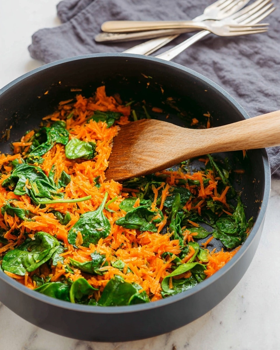 A black pan is filled with two main layers of food, the bottom layer is bright orange shredded carrots, and fresh green spinach leaves are spread on top. A wooden spoon is stirring the mix, which looks fresh and lightly cooked, with some spinach leaves lying over the carrots and others slightly mixed in. The pan rests on a white marbled surface. In the background, there is a gray cloth with three silver and wooden-handled forks placed on it. Photo taken with an iphone --ar 4:5 --v 7
