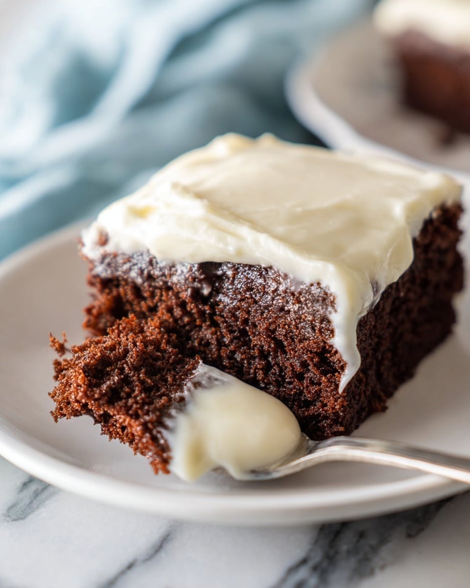 A close-up image of a single square piece of chocolate cake on a white plate, showing two layers: the bottom layer is a rich, dark brown cake with a soft, slightly crumbly texture, and the top layer is smooth, creamy white frosting spread evenly across the cake's surface. A spoon holds a small portion of the cake with a thick layer of frosting dripping slightly, highlighting the contrast of the dark chocolate cake and the light frosting. The background features a white marbled texture under the plate and a soft focus on a light blue cloth underneath. Photo taken with an iphone --ar 4:5 --v 7