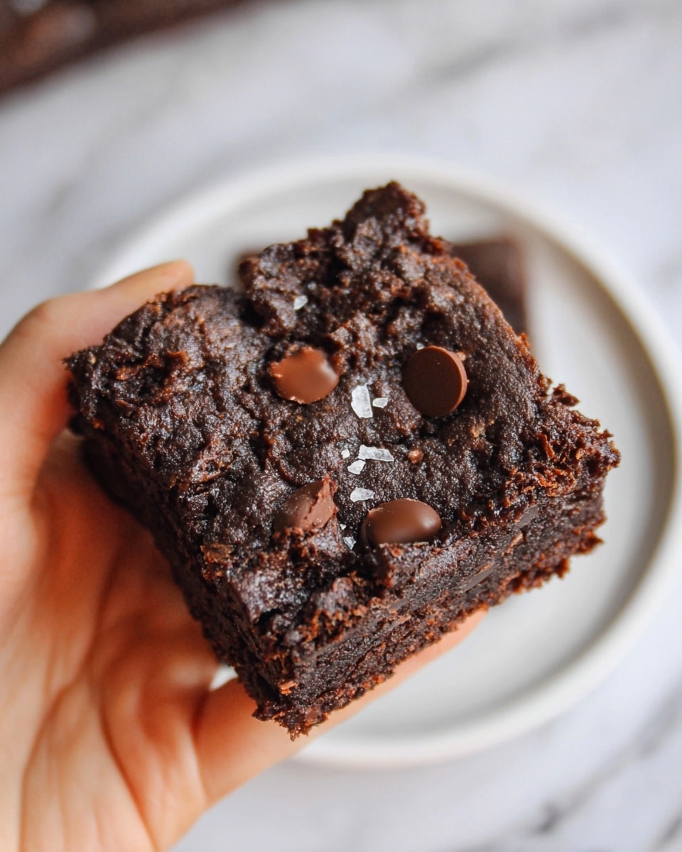 A close-up image shows a single square chocolate brownie held by a woman's hand. The brownie is dark brown with a rough, textured surface. It has three visible chocolate chips on top, slightly melting into the brownie, and small bits of salt sprinkled over the top. The background is a white marbled texture, and there is a white plate visible in the background. The brownie looks dense and rich with a slightly shiny finish on the edges. Photo taken with an iphone --ar 4:5 --v 7