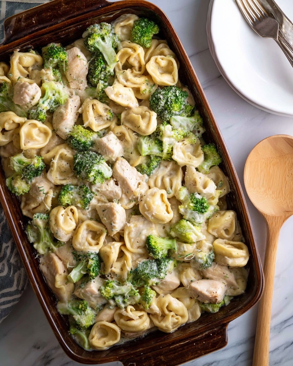 The dish shows a close-up of a creamy pasta mix in a dark baking dish, filled with three main layers: light beige tortellini pasta shaped in small rings with soft folds, chunks of lightly browned chicken pieces that have a smooth texture, and vibrant green broccoli florets with a slightly rough surface. All ingredients are coated in a smooth, creamy white sauce that ties the elements together. The dark baking dish contrasts with the bright food, all placed on a white marbled surface. photo taken with an iphone --ar 4:5 --v 7