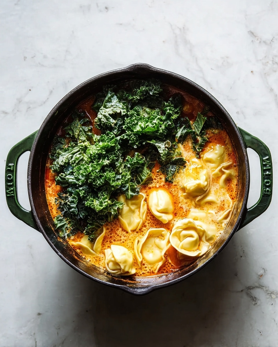 A dark green pot filled with three main layers: on the left side, fresh dark green kale leaves with a rough texture; on the right side, pale yellow tortellini pasta with a smooth, folded shape; and underneath these, an orange-red broth with a slightly creamy surface showing some bubbles. The pot sits on a white marbled surface. Photo taken with an iphone --ar 4:5 --v 7