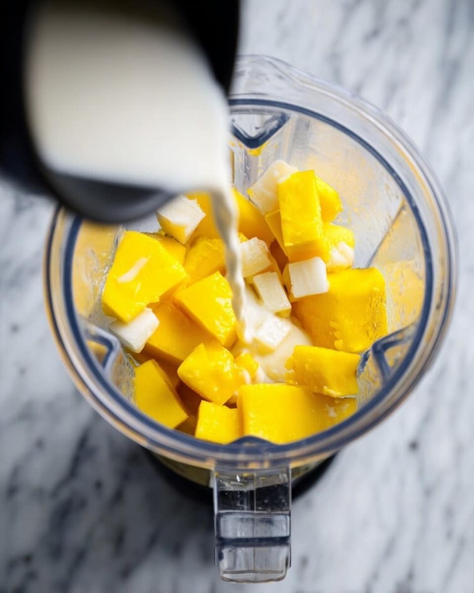 A clear blender container filled with chunky pieces of bright yellow mango and small white cubes, with a black cup pouring white liquid over the mango and cubes inside the blender, all set on a white marbled surface photo taken with an iphone --ar 4:5 --v 7