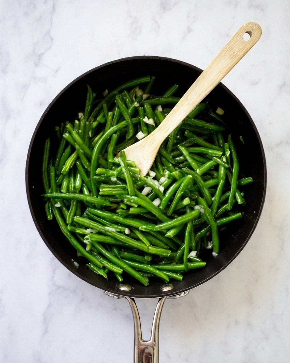 A black pan filled with bright green cut green beans, showing a fresh and slightly shiny texture. The green beans are evenly spread, with some small pieces of chopped white onions mixed in. A long, light-colored wooden spoon rests inside the pan, lying diagonally from the bottom right to the top center, slightly stirring the beans. The pan handle is metal and extends upward, placed on a white marbled surface. The whole scene looks clean and simple, with a top-down view photo taken with an iphone --ar 4:5 --v 7