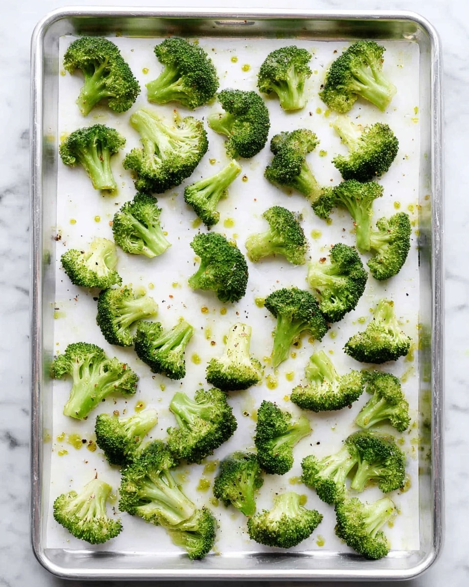 The image shows a single layer of broccoli pieces spread out evenly on a baking tray lined with white parchment paper. The broccoli is bright green with firm stalks and bushy tops, some pieces slightly different in size and shape, with a few drops of oil and small specks of seasoning scattered across the paper. The tray edges are silver, and the background surface has a white marbled texture. Photo taken with an iphone --ar 4:5 --v 7