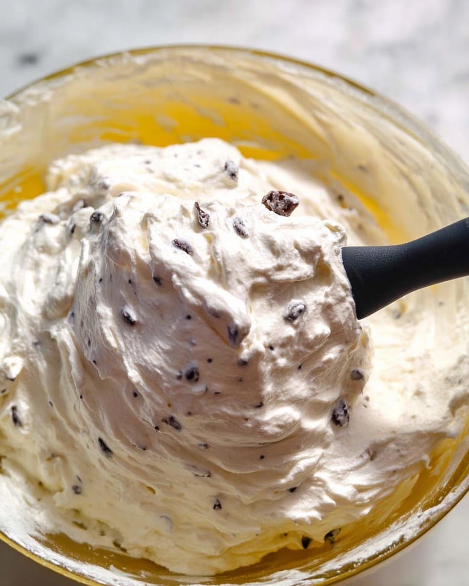 A close-up view of thick white cream mixed with small dark chocolate chips, showing a fluffy and smooth texture with slight peaks. The cream is in a clear glass bowl, with the yellowish inside of the bowl slightly visible. A black spatula with a matte handle is scooping some of the creamy mixture, highlighting the texture and the chocolate bits embedded inside. The bowl is placed on a white marbled surface, creating a clean and bright background. photo taken with an iphone --ar 4:5 --v 7