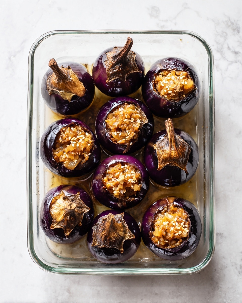 The image shows a clear rectangular glass baking dish on a white marbled surface holding eight roasted small eggplants arranged in two rows. Each eggplant is dark purple, with split and wrinkled shiny skin revealing soft, light yellowish insides. The eggplants have brown dried stems on top. In the upper part of the image, there is a round white bowl containing a chunky mixture of cooked ingredients in shades of light brown and beige, with small bits that look like sesame seeds or grains scattered on top. The overall scene is clean and bright. photo taken with an iphone --ar 4:5 --v 7