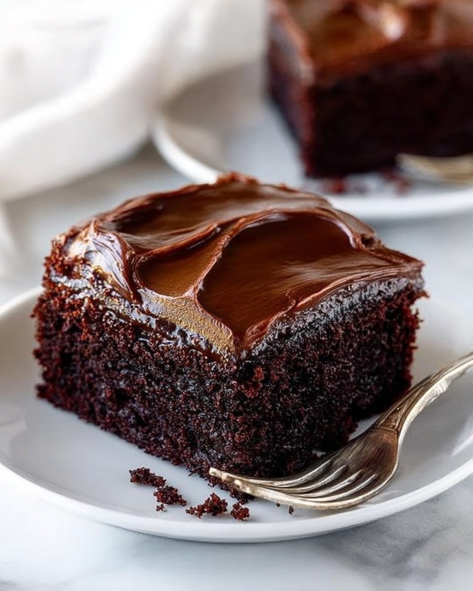 A square piece of dark chocolate cake sits in the center of a white plate on a white marbled surface, topped with a smooth, thick layer of glossy chocolate frosting that is slightly swirled on top. The cake layer is rich and moist with a dense texture, showing tiny crumbs around the edges. A silver fork rests on the right side of the plate. In the background, another similar piece of cake on a white plate is slightly out of focus. Photo taken with an iphone --ar 4:5 --v 7