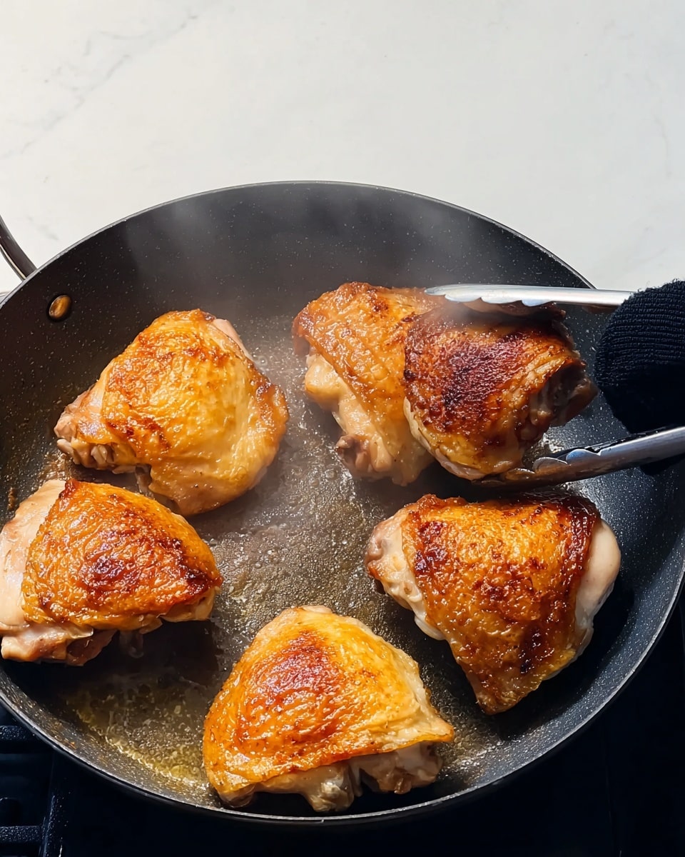 Five pieces of chicken with golden brown, crispy skin are cooking in a dark frying pan. The chicken pieces have a slightly raised texture on the skin and look juicy underneath. A woman's hand wearing a black silicone oven mitt is holding one piece of chicken with tongs on the right side of the pan. The pan sits on a white marbled countertop with a plain light background. Steam rises gently from the chicken showing that it is hot. photo taken with an iphone --ar 4:5 --v 7