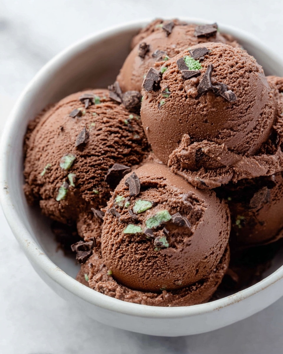 A close-up view of several scoops of rich chocolate ice cream with a smooth but slightly rough texture, piled on top of each other. The ice cream is dark brown with small green bits mixed in, and sprinkled with pieces of dark chocolate on the top layer. The scoops sit inside a white bowl, which is set on a white marbled surface. Photo taken with an iphone --ar 4:5 --v 7