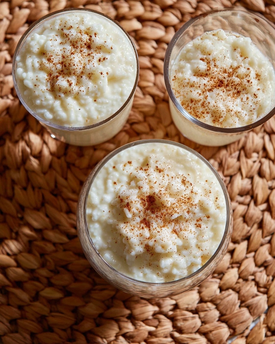 The image shows one large transparent glass bowl and two smaller transparent glasses filled with creamy rice pudding. Each container is filled to the top with thick, textured white rice pudding that has visible small rice grains. The pudding is sprinkled lightly with a brown spice, likely cinnamon, adding contrast on the surface of each. The containers are placed on a woven, natural-colored textured mat with a warm tone. The scene is viewed from above with a clear focus on the creamy texture and spice topping of the rice pudding. photo taken with an iphone --ar 4:5 --v 7