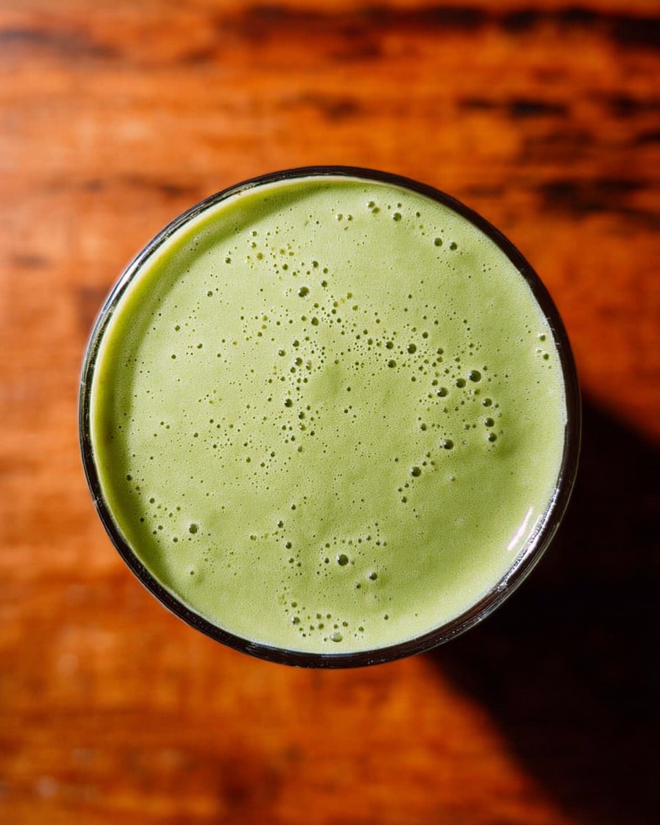 The image shows a top view of a single glass filled with a smooth, light green smoothie. The surface of the smoothie is creamy with small bubbles scattered across it, creating a slightly frothy texture. The glass is round and the green smoothie fills it almost to the edge. The background is a warm-toned wooden texture that has been changed to a white marbled texture. The photo taken with an iphone --ar 4:5 --v 7