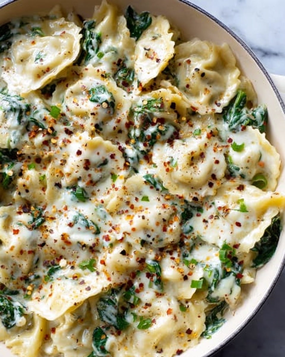 The image shows a white pan filled with pasta layered with melted white cheese and green spinach mixed evenly throughout. The surface of the cheese is slightly browned and sprinkled with red pepper flakes and black pepper for added texture and color contrast. The pasta shapes are mostly half-moon ravioli pieces, clearly visible under the cheese, and the green spinach leaves are scattered in between adding a fresh look. The background is a white marbled texture. photo taken with an iphone --ar 4:5 --v 7