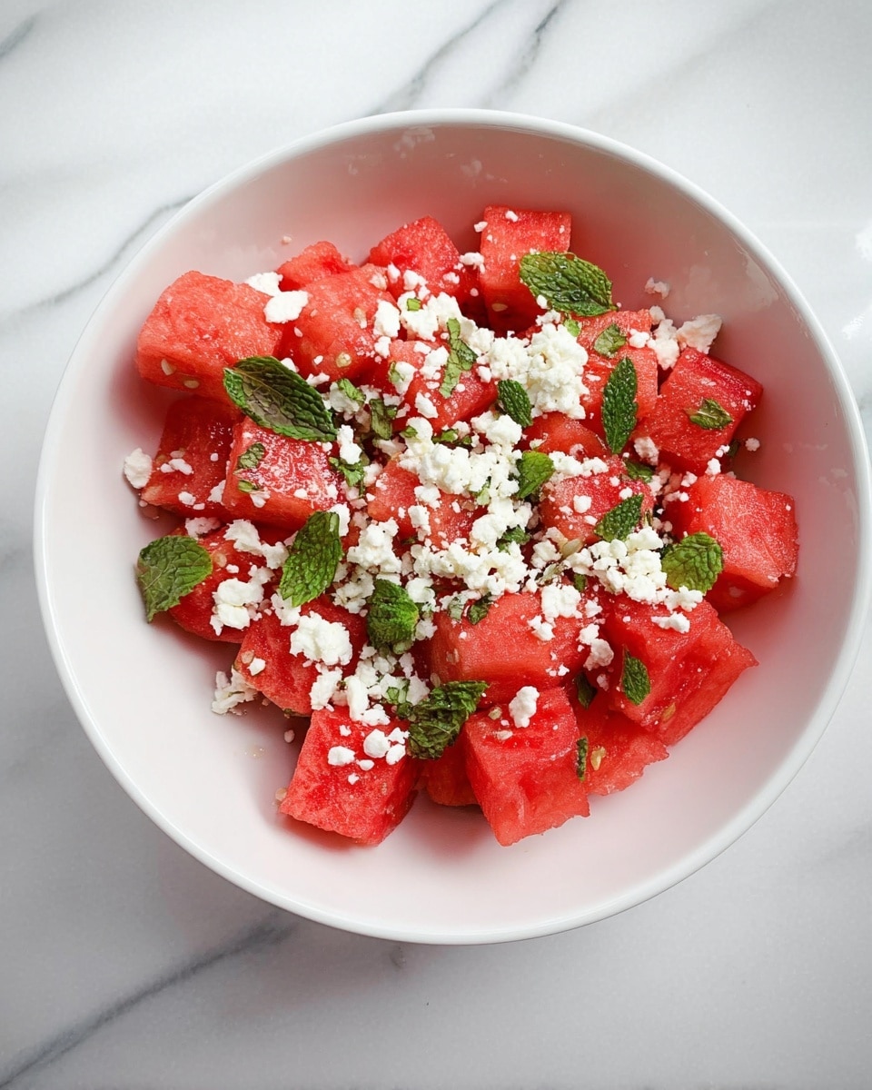 A white bowl filled with several chunks of bright red watermelon as the bottom layer, scattered with small pieces of fresh green mint leaves. On top, there is a layer of small, crumbly white cheese bits spread evenly over the watermelon. The bowl is placed on a white marbled surface. photo taken with an iphone --ar 4:5 --v 7