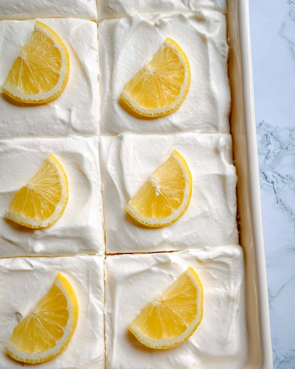 A close-up view of a large rectangular dessert with one smooth layer of white cream evenly spread on top. The dessert is divided into neat square sections, each topped with a fresh yellow lemon wedge placed near the center. The dessert sits inside a white pan with soft, creamy texture visible at the edges. The background shows a white marbled surface with subtle natural patterns. photo taken with an iphone --ar 4:5 --v 7
