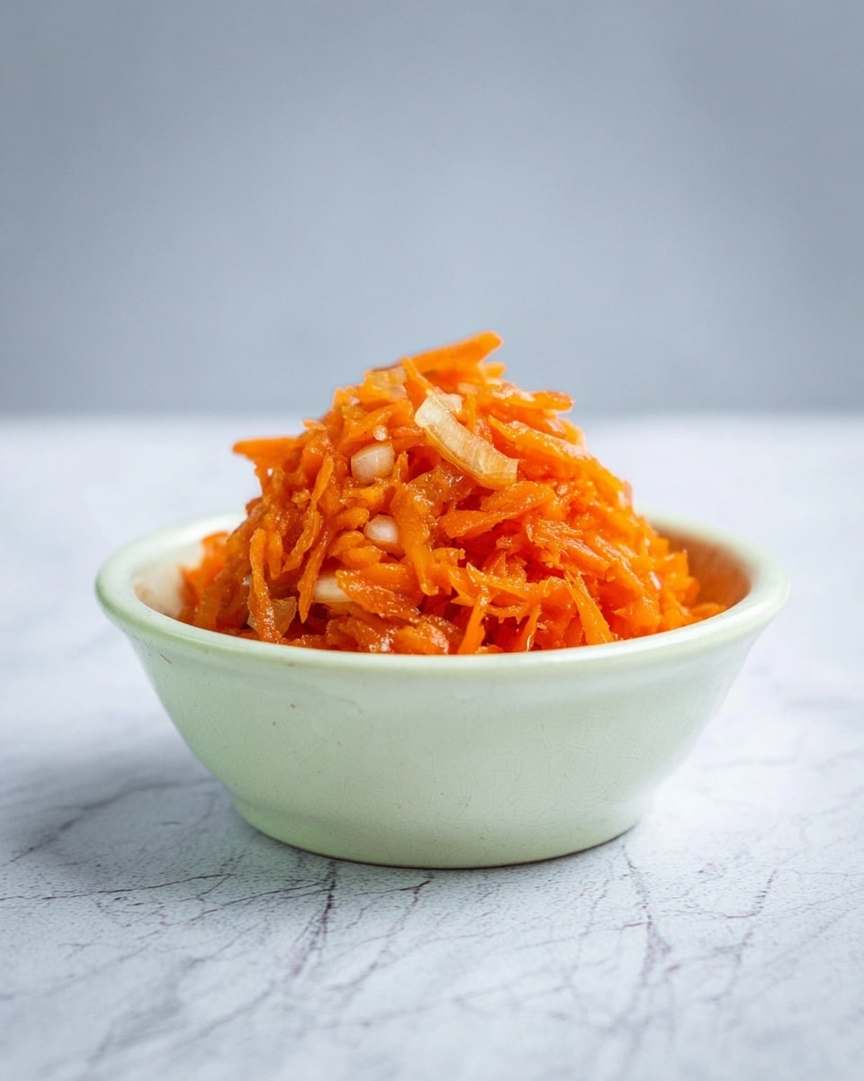 A small white bowl filled with a mound of shredded carrot salad that has a shiny, slightly wet texture showing thin pieces of orange carrot mixed with small bits of light-colored onions or similar vegetables, placed on a white marbled surface with a simple light gray background, photo taken with an iphone --ar 4:5 --v 7