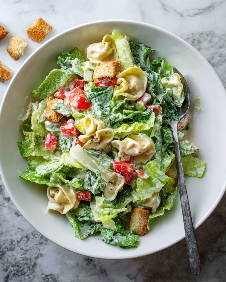 A white bowl filled with a fresh salad showing three main layers: green romaine lettuce leaves with a creamy white dressing spread unevenly, small red diced tomato pieces scattered mostly on one side, and pale-yellow folded tortellini pasta mixed throughout. Small light brown croutons are spread on the salad and some lie outside the bowl on a white marbled surface. A silver fork rests in the salad near the bottom right inside the bowl. Photo taken with an iphone --ar 4:5 --v 7