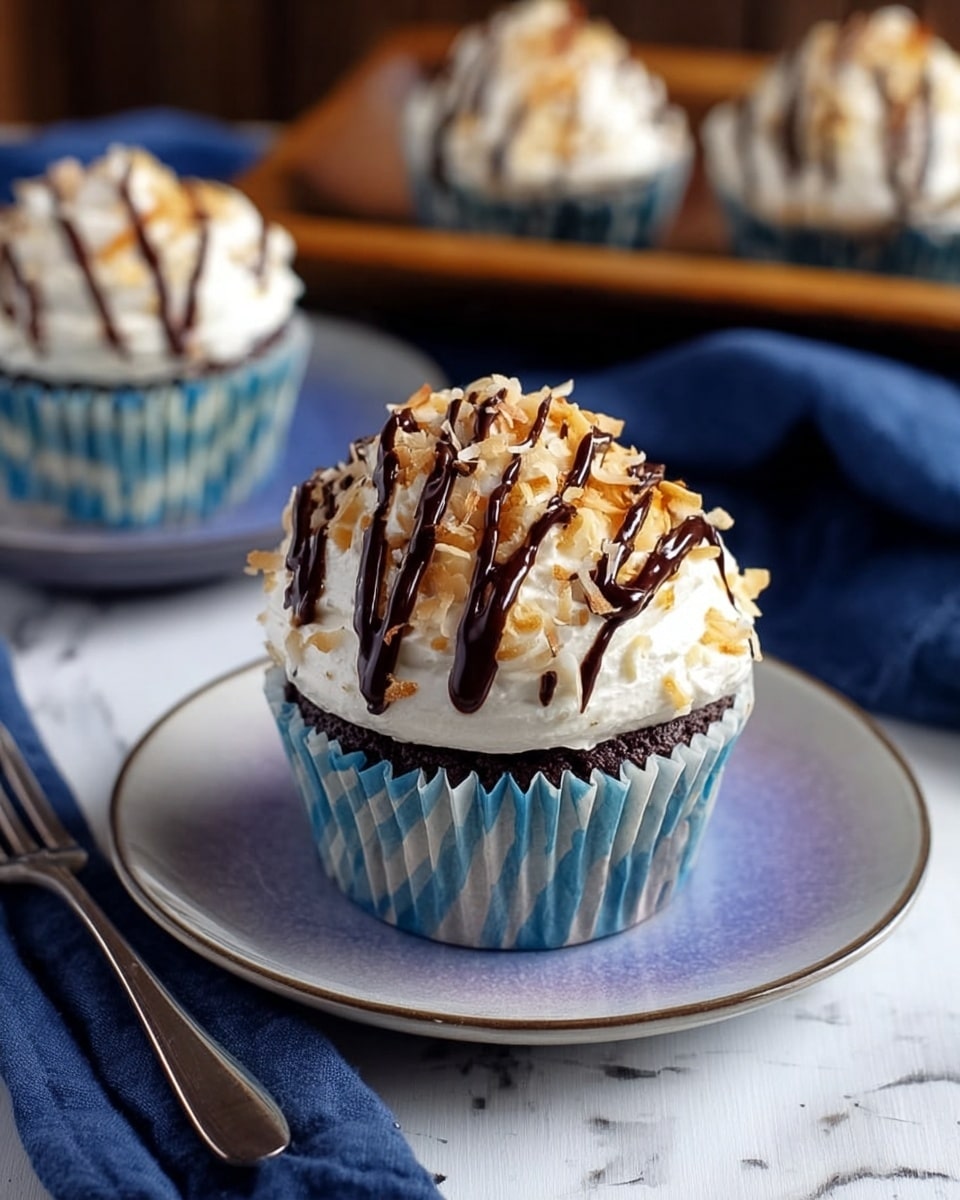 A single cupcake sits on a white plate with a bluish-purple tint, placed on a white marbled surface next to a silver fork and a dark blue cloth napkin. The cupcake has a chocolate base in a blue-striped paper liner. It is topped with a thick layer of white, fluffy frosting sprinkled with toasted coconut flakes and drizzled with dark chocolate stripes across the top. In the background, there is a wooden tray with more cupcakes on it. The photo taken with an iphone --ar 4:5 --v 7